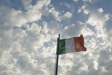 background: Italian flag, green, white, red, waving in the air on an afternoon of strong wind, blue sky full of white clouds, by the sea of ??Liguria, Italy