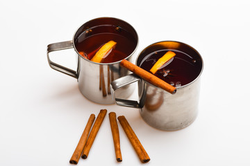 Mulled wine or beverage with cinnamon. Mugs with mulled wine or hot drink and cinnamon aroma sticks, white background. Hot beverage concept. Tea or hot drink in metal mug with slise of orange