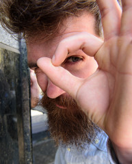 Watching concept. Guy looks suspicious through ok gesture, close up. Hipster with tousled hair looking, observing, watching. Man with beard on thoughtful, suspicious face, black marble background.