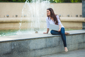 beautiful business girl brunette sitting with laptop at a fountain in the street