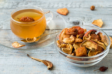 A mixture of dried fruits in a small round glass bowl, next to a mug with compote on an old table.