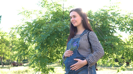 A young, beautiful, pregnant girl, near a tree, dressed in overalls.