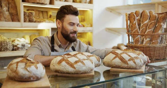 Portrait of the young handsome male baker standing and leaning on the counter in the bakery shop while smelling fresh bread. Indoor
