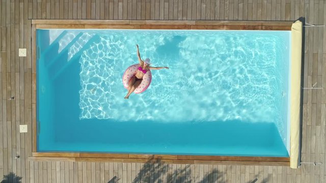 AERIAL, TOP DOWN, CINEMAGRAPH: Joyful Girl In Pink Swimsuit Chilling On A Doughnut Floatie In The Summer. Gorgeous Woman Suntans While Floating Around The Crystal Clear Pool On Inflatable Doughnut.