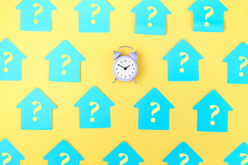 On a yellow background, many blue stickers in the form of a house with question marks are attached. In the center is a small alarm clock. Concept, a matter of time.