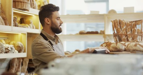 Young handsome man vendor in the apron bringing fresh bread to the counter in the bakery shop. Indoors