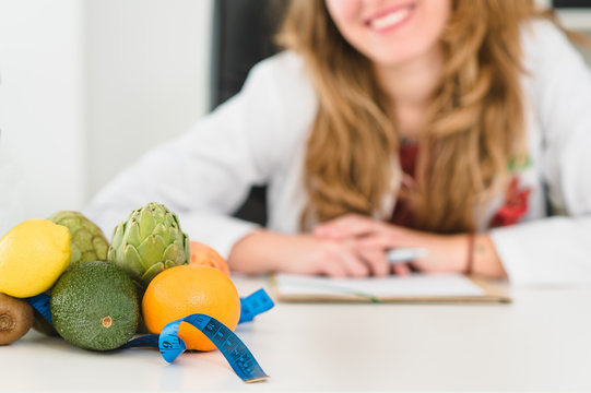 Nutricionista Caucásica Sonriendo En La Clínica Con Una Bata Blanca Y Fruta En El Escritorio. Comida Sana Con Un Metro Medidor . Naranjas, Alcachofas, Aguacate Y Limones 