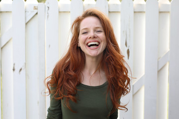 Hübsche rothaarige Frau vor weißem Zaun lacht begeistert
