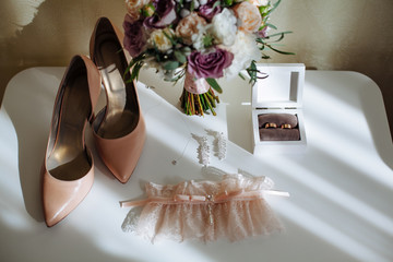 Bridal morning details composition. Top view of wedding rings, beautiful bouquet of violet flowers with ribbons, earrings, garter and leather shoes. Flat lay.