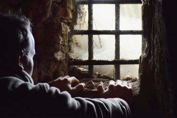 Man prisoner watching out from underground window, hands on the wall