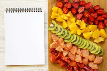 Chopped fresh colorful fruits arranged on cutting board with notepad on white wooden background, closeup. Top view.