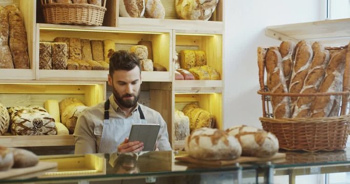 Caucasian man bread seller holding his tablet computer in hands while standing at the counter in the bakery shop and looking at the side in the window. Indoor
