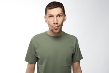 Stylish unshaven man over studio wall makes grimace. Portrait of bearded young male wearing trendy t-shirt rounds lips, has trendy hairdo and posing at camera isolated. Copy space for advertising.