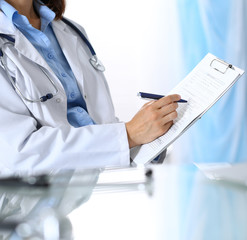 Female doctor filling up medical form on clipboard, closeup. Reflecting glass table is a physician working place. Healthcare, insurance and medicine concept