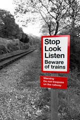 Railway warning sign in black and white with red signpost reading "Stop Look Listen Beware of trains" with a railway running off into the distance.