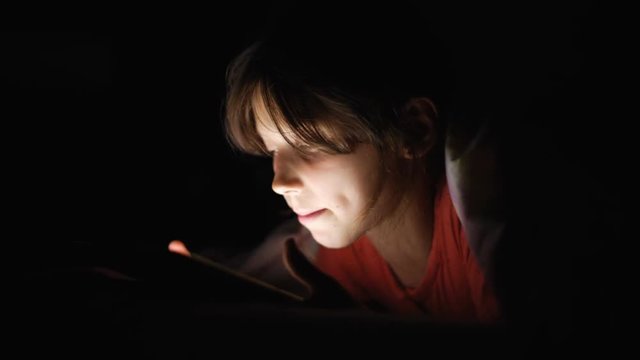 Dolly shot caucasian girl lying in bed playing tablet in social internet in dark light under blacket. Childhood, communication and technology concept