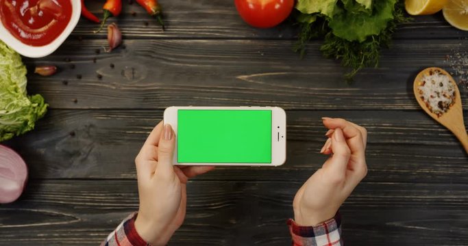 View from above on the white horizontal smart phone on the dark wooden table with food background and female hands scrolling, taping and zooming on its green screen. Chroma key. Close up