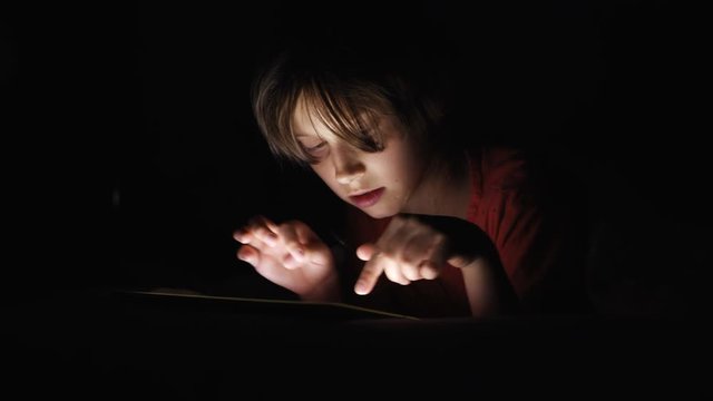 Dolly shot caucasian girl lying in bed playing tablet in social internet in dark light under blacket. Childhood, communication and technology concept