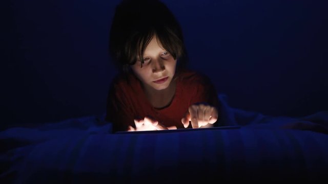 Dolly shot caucasian girl lying in bed playing tablet in social internet in dark blue light under blacket. Childhood, communication and technology concept