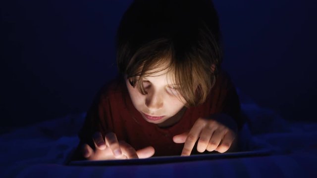 Dolly shot caucasian girl lying in bed playing tablet in social internet in dark blue light under blacket. Childhood, communication and technology concept