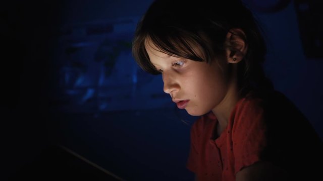 Dolly shot caucasian girl lying in bed playing tablet in social internet in dark blue light under blacket. Childhood, communication and technology concept