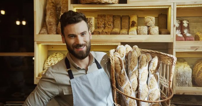 Portrait of the handsome young male baker smiling while posing with baguettes in the basket in the shop. Indoors
