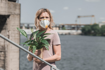 woman in protective mask holding potted plant on bridge, air pollution concept