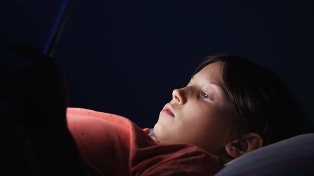 Caucasian girl lying in bed playing tablet in social internet in dark blue light under blacket. Childhood, communication and technology concept