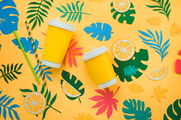 Lemonade paper cups in tropical header with oranges, leaves and fruits pattern on a bright yellow background. Sunny summer colorful flat lay with paper cut outs.