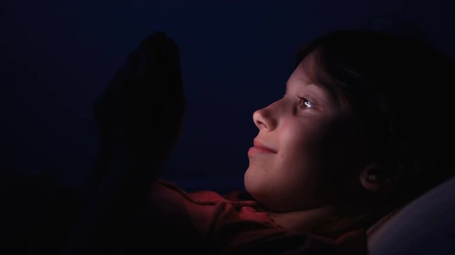 Dolly shot caucasian girl lying in bed playing tablet in social internet in dark blue light under blacket. Childhood, communication and technology concept