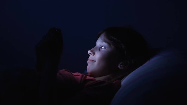 Dolly shot caucasian girl lying in bed playing tablet in social internet in dark blue light under blacket. Childhood, communication and technology concept