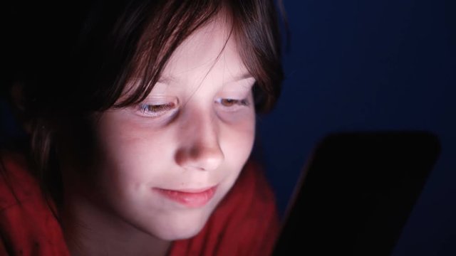 Caucasian girl lying in bed playing tablet in social internet in dark blue light under blacket. Childhood, communication and technology concept
