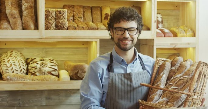 Portrait shot of the handsome male baker smiling while posing with baguettes in the basket in the shop. Inside