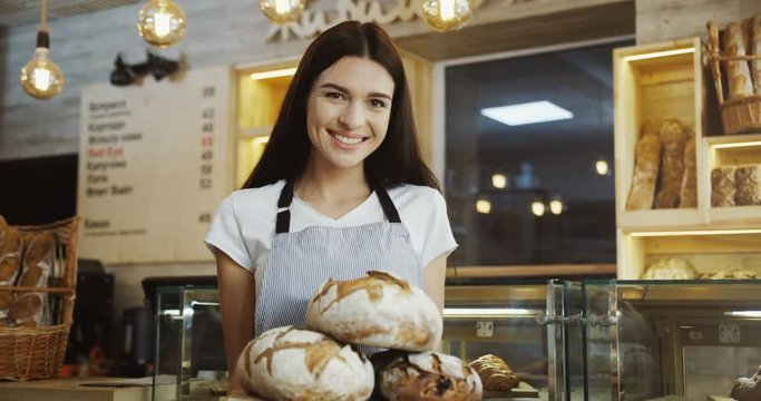 Portrait of the caucasian young woman baker looking at the camera while holding a tray with bread in the shop. Indoors