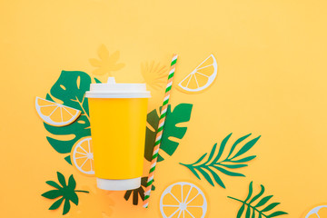 Disposable paper cup on a yellow background with tropical leaves and orange slices on a bright yellow background. Colorful summer drink concept with paper cut outs.