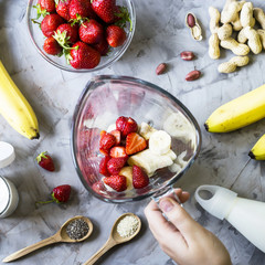 Ingredients for making strawberry banana smoothies on a gray table next to a glass bowl of blendet. Cooking a healthy breakfast concept. TOp view, flat lay