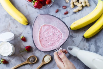 Ingredients for making strawberry banana smoothies on a gray table next to a glass bowl of blendet. Cooking a healthy breakfast concept. TOp view, flat lay