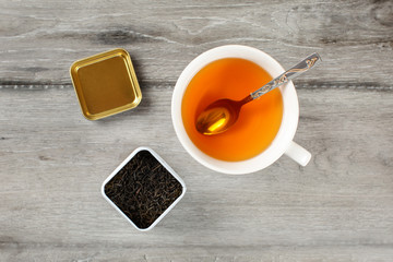 Table top view on cup of hot amber tea with spoon, and caddy full of black dried loose leaves,...