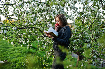 Young brunette girl at jeans against spring blossom tree and read the book and eat apple.