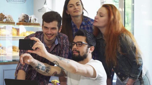 Young bearded handsome man using his smart phone taking selfies with a group of his friends at the cafe. Cheerful friends taking photos together. Relationships, social media, technology, lifestyle.