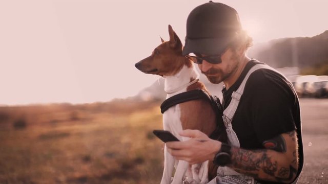 Millennial hipster stylish man with authentic tattoos sits on seaside at sunset with best friend puppy dog on lap, texts or uses smartphone application. concept connectivity or communication