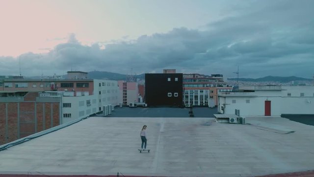 Epic and inspiring drone aerial footage of cool and trendy hipster millennial teenager or skater woman skate on top of rooftop on urban city building at astonishing sunset, teen lifestyle