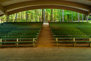 Empty benches and an empty stage on this outdoor arena in a public park. Trees and parts of houses visible in the background.