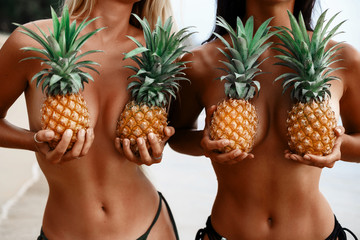 two incredibly beautiful sexy girl models in a bikini on the beach of a tropical island, blond brunette, bronze tan, travel summer vacation, fashion style, in the hands of pineapples cover the chest - obrazy, fototapety, plakaty