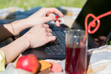 Female hands work on the laptop keyboard on a picnic. A freelancer girl is working with a laptop on the grass in the park. Nearby are a glass of juice and a plate with biscuits.