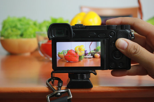 Photographer taking photo of fresh fruits and vegetables on camera display while shooting in kitchen table