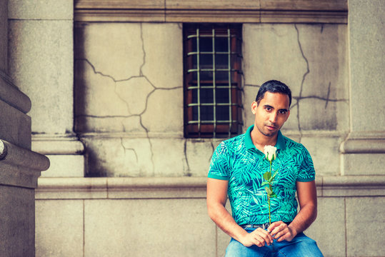 Young Hispanic American Man Missing You, Waiting For You, Wearing Green Patterned Polo Shirt, Holding White Rose, Sitting On Old Street In New York, Looking Forward..