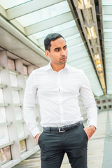 Portrait of Young Handsome Hispanic American Man in New York, wearing white shirt, black pants, standing on walkway with glass wall inside office building, looking down, thinking..