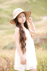Cute kid girl wearing white dress and hat stading in meadow. Childhood. Summer time.