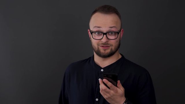 A young, bearded man with glasses. Studio shot of male looks at his phone in amazement. Watches and can not recover. Emotionally reacts to the message.